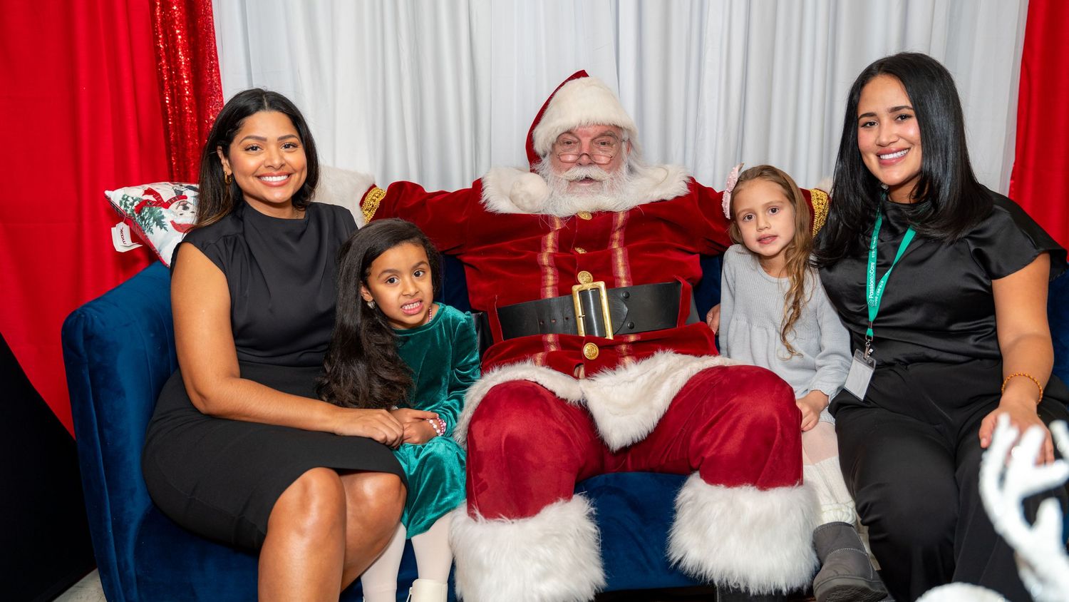 Traditional holiday scene with Santa Claus meeting families in front of red and white backdrop.