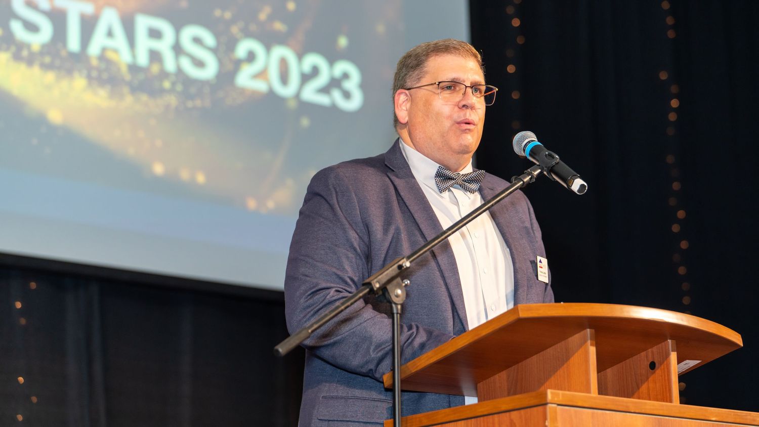 Speaker presents at a podium during a 2023 event with illuminated stage backdrop.
