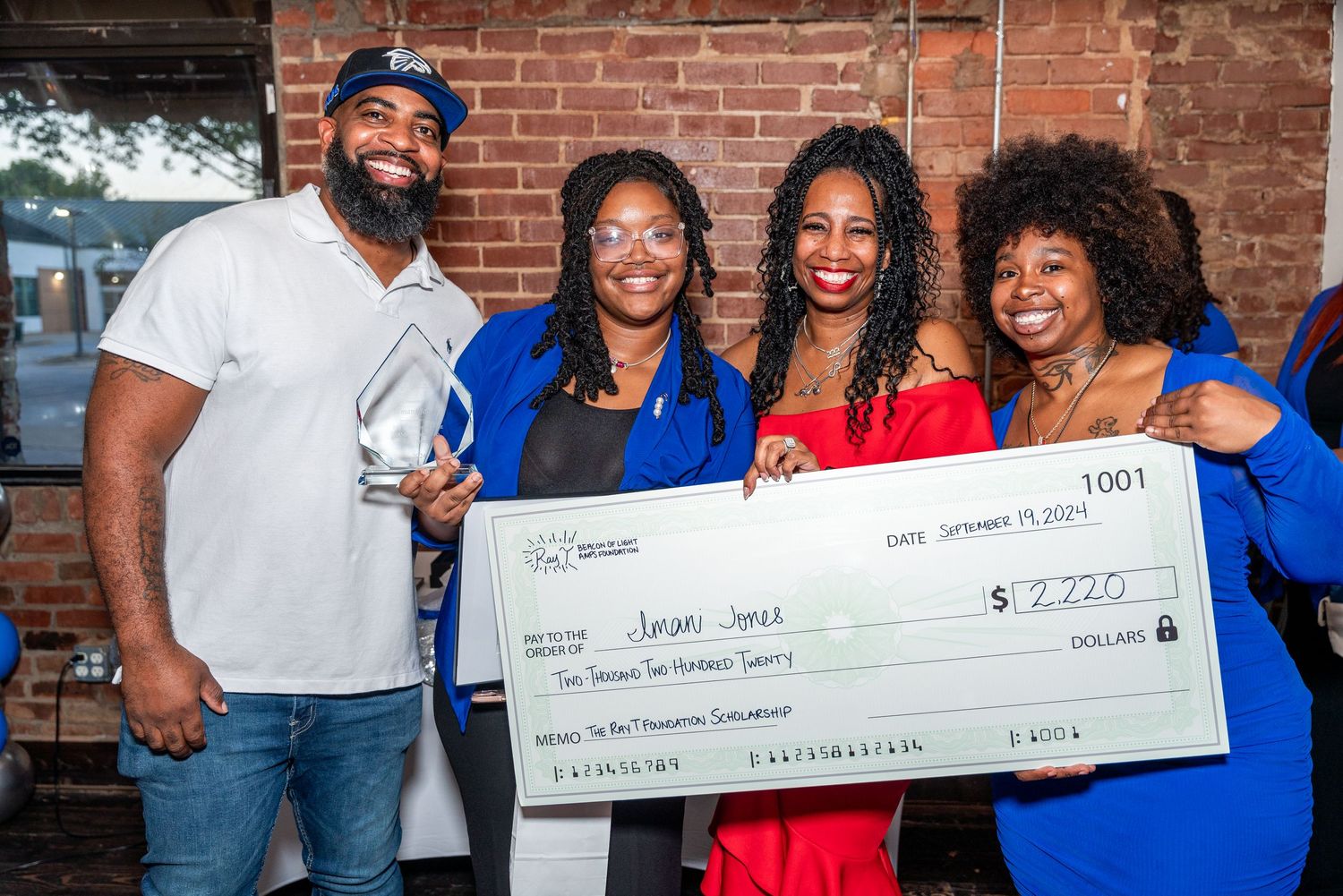 Group celebrates with large check presentation against brick wall backdrop.