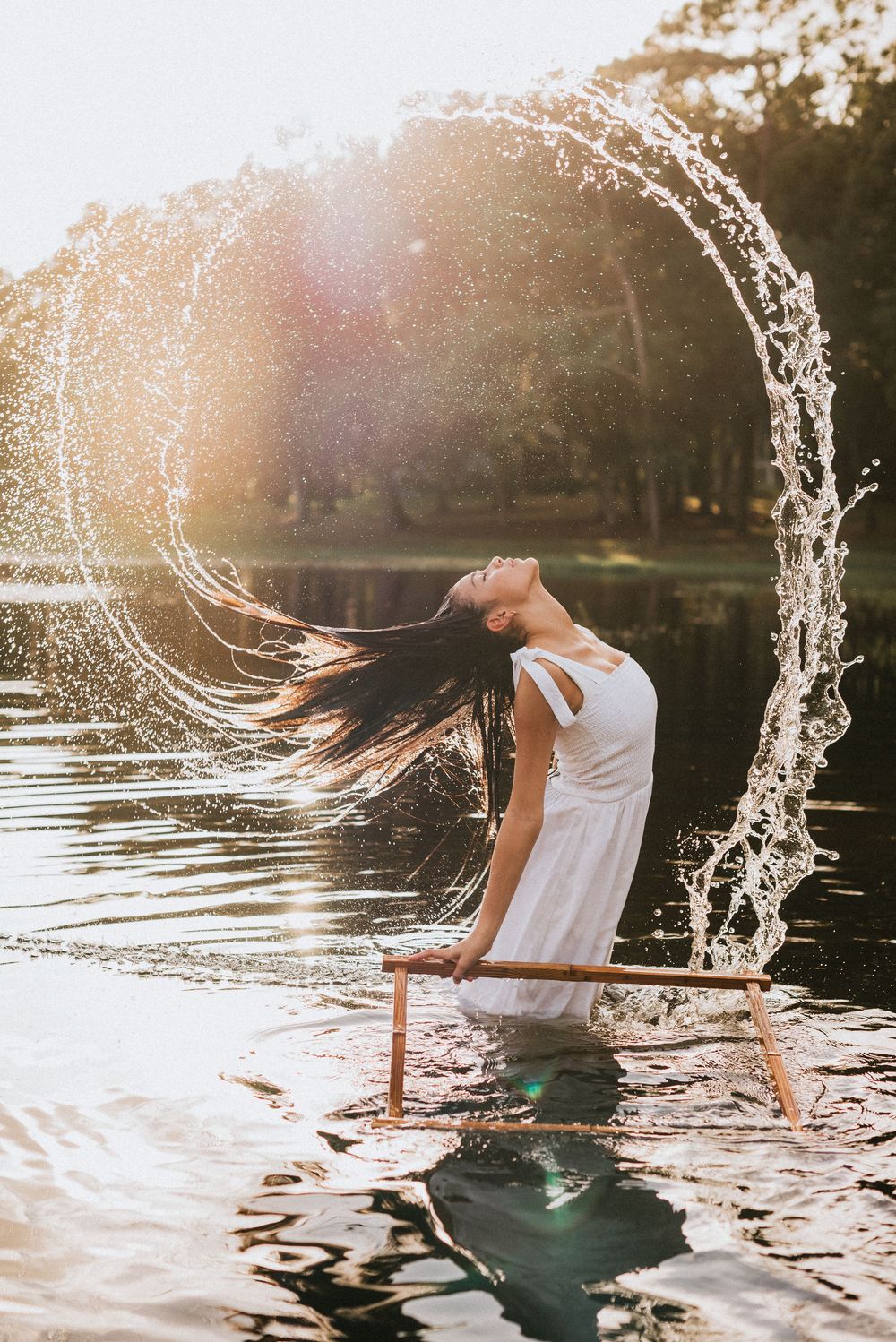Woman in white dress flipping wet hair creating arc water splash while standing in sunlit lake during a Lake Mary photo shoot.