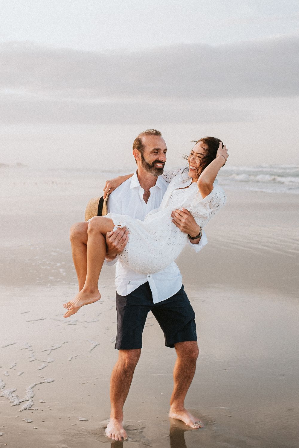 At new smyrna beach a man carries a woman in his arms on a sandy beach, both smiling and wearing white, with ocean waves behind them.