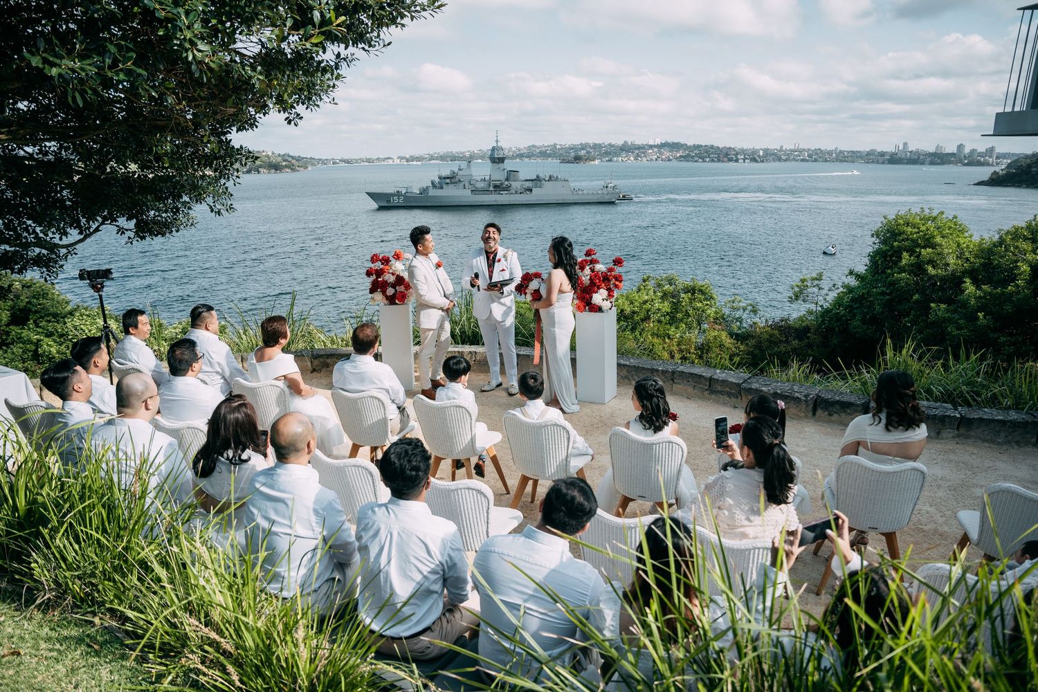 Waterfront wedding ceremony with guests seated on hillside overlooking harbor.