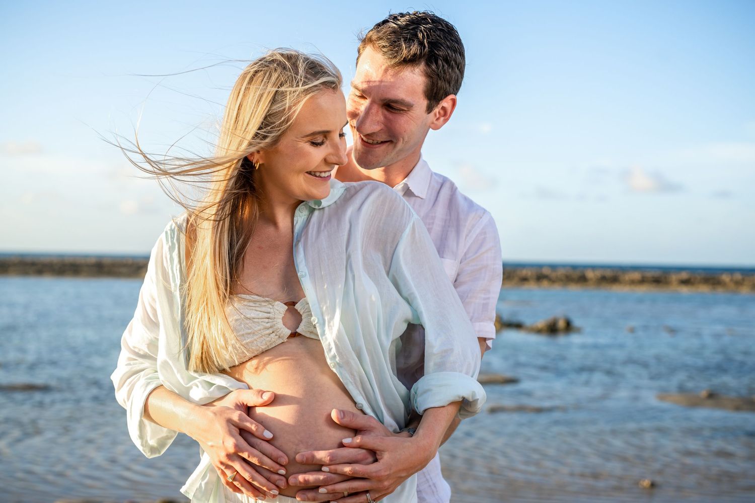 Intimate beachside maternity photo with couple embracing near ocean rocks.