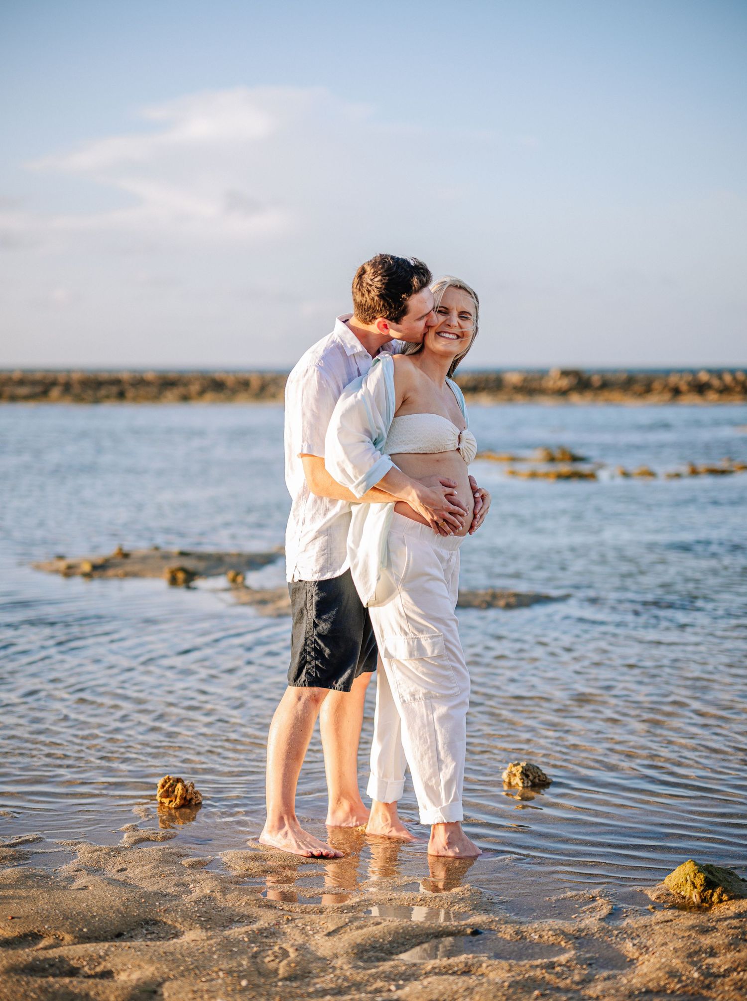 Joyful couple shares tender moment during beach maternity photoshoot.