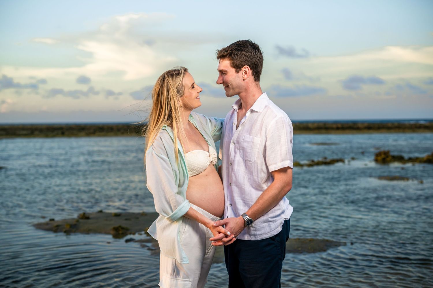 Expecting couple in light clothing shares intimate moment at oceanside.