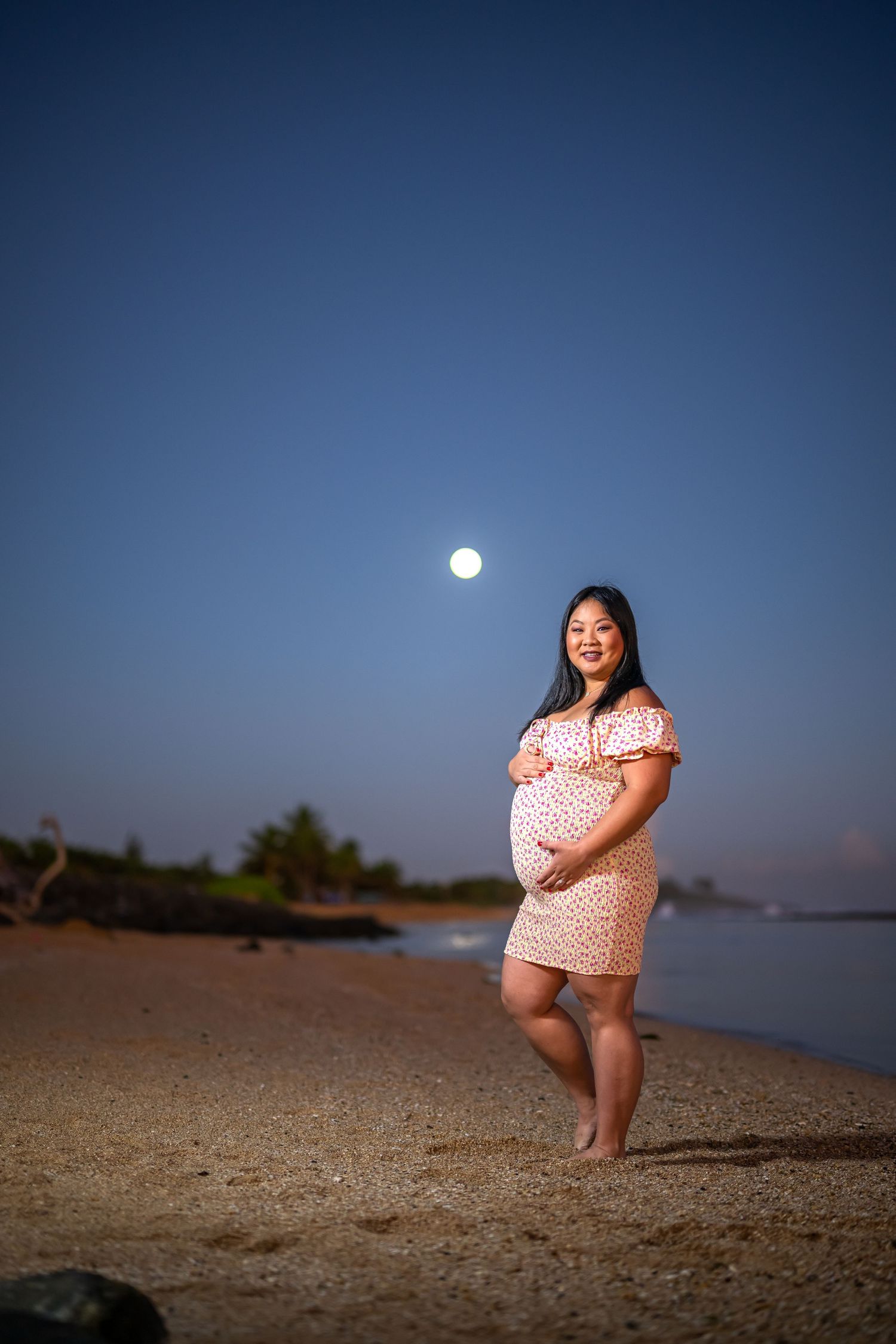 Nighttime maternity portrait on beach with full moon and palm trees in background.