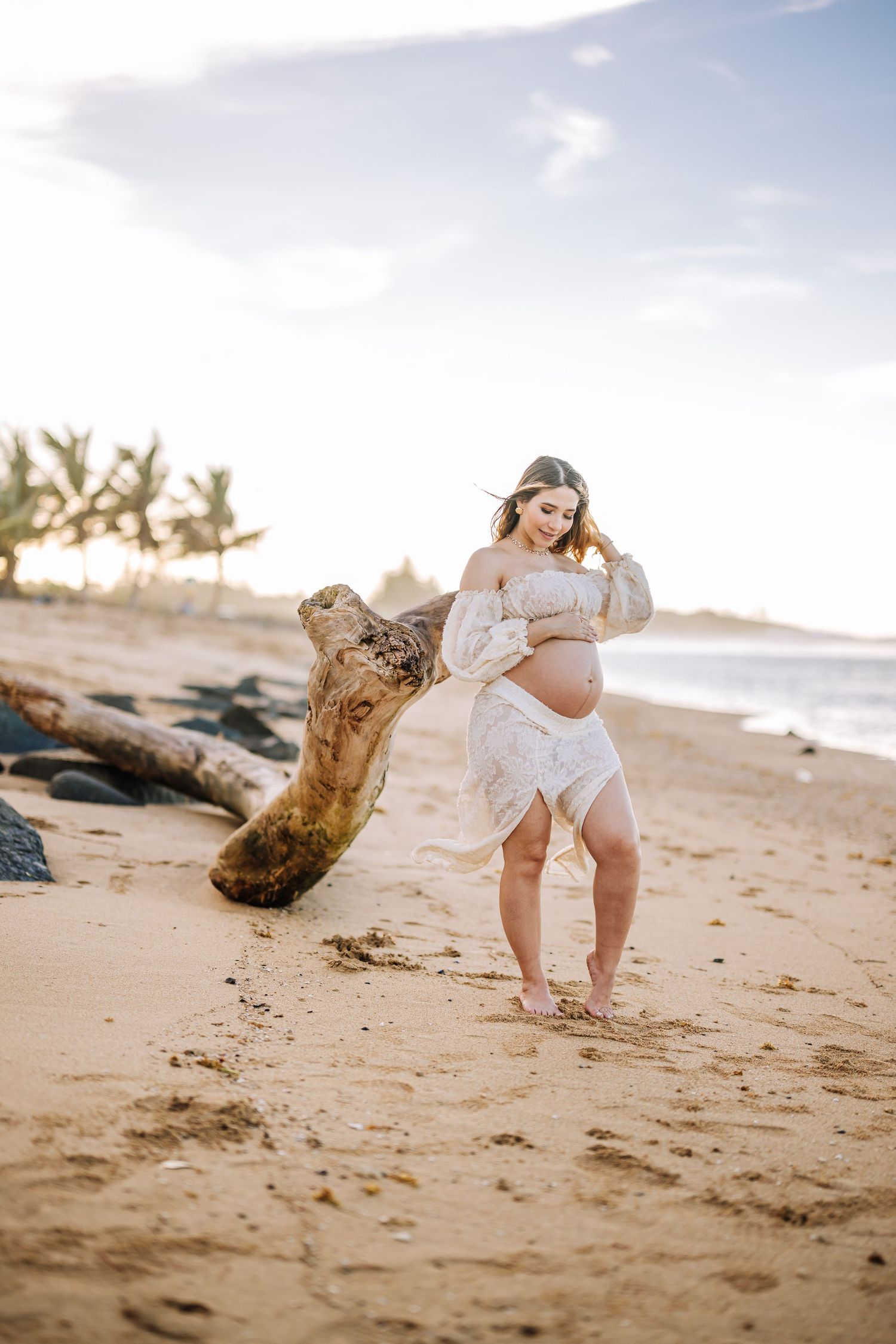 Expecting mother in white dress poses on sandy beach with driftwood at golden hour.