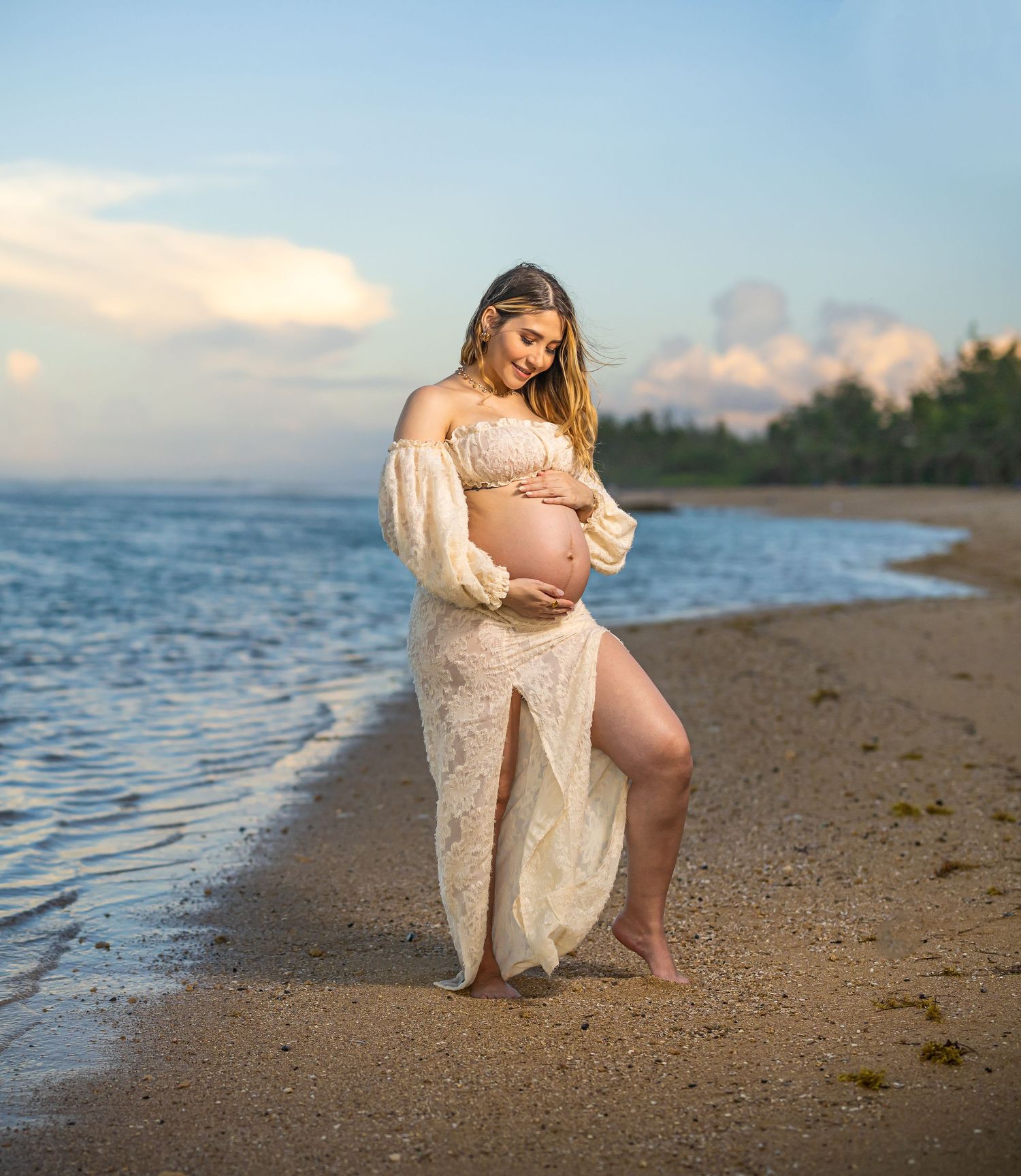 Maternity beach photo with flowing cream dress against ocean backdrop at sunset.
