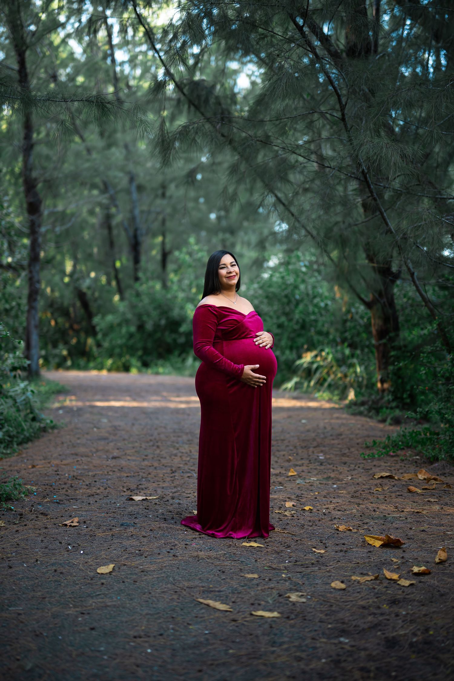 Maternity portrait in a burgundy gown along a forest path surrounded by lush greenery.