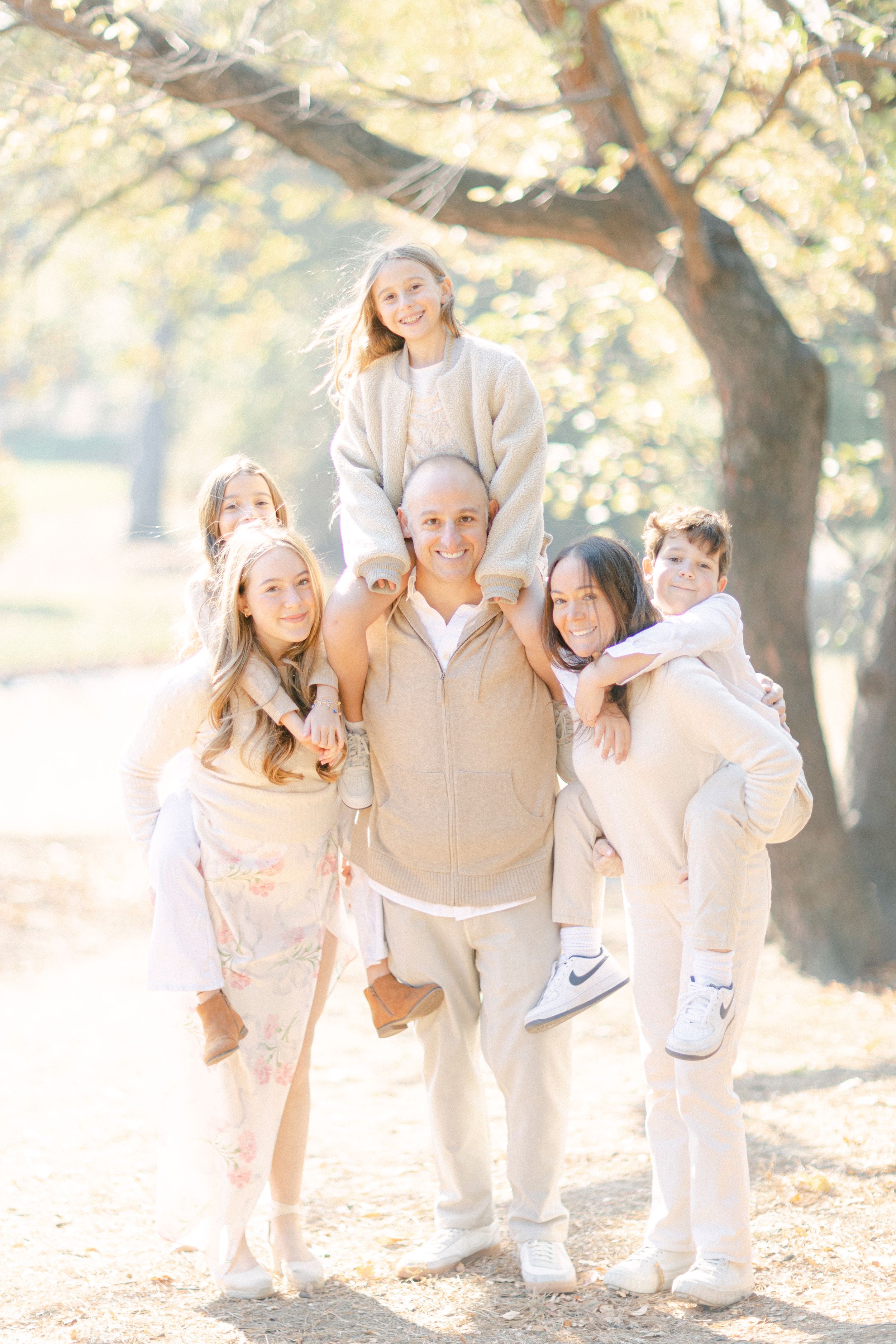 Family group in coordinated light-colored outfits poses together under blooming trees in a park.