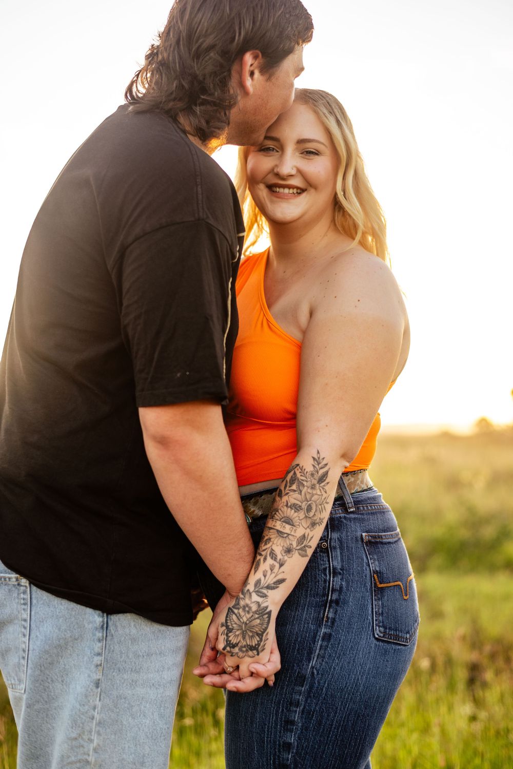 A romantic couple embraces in golden sunset light, showing affection while standing in a grassy field.