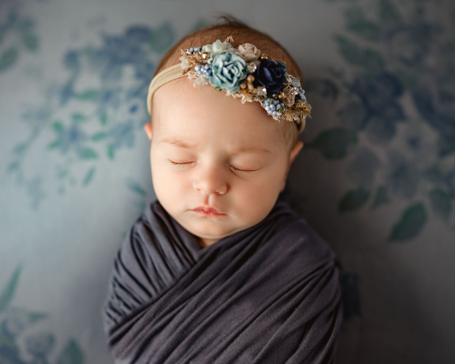 Sleeping newborn wrapped in gray swaddle with delicate floral headband against blue floral backdrop. Middle TN Newborn Photographer.