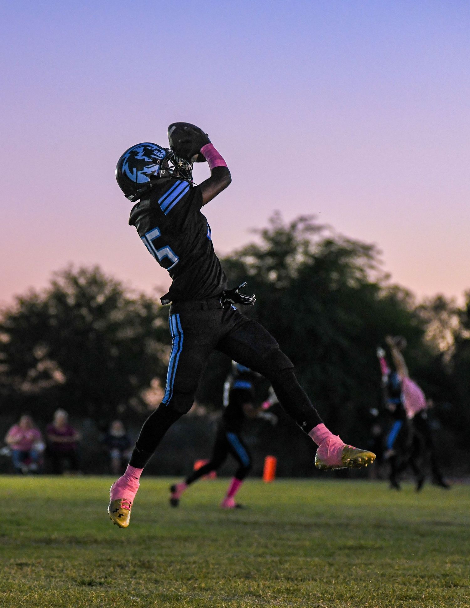 Football player in black and blue uniform jumping to catch the ball during a sunset game.