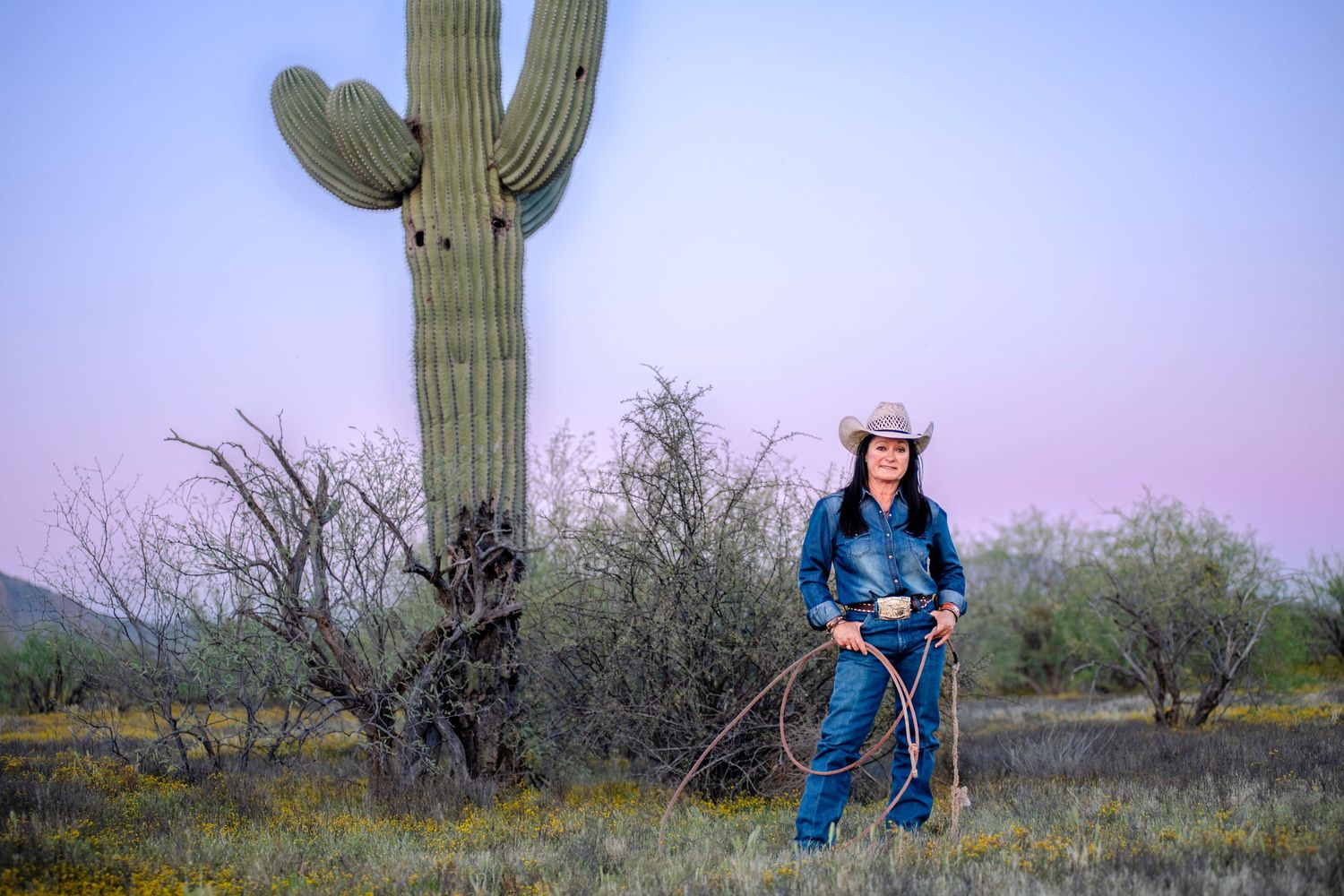 Cowgirl posing with lasso near a towering saguaro cactus during purple desert sunset.