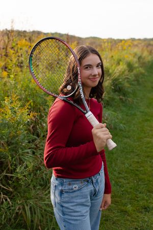 Athletic person holds tennis racquet while wearing red sweater and jeans in an autumn outdoor setting. Senior photo session. Lake Geneva Wis