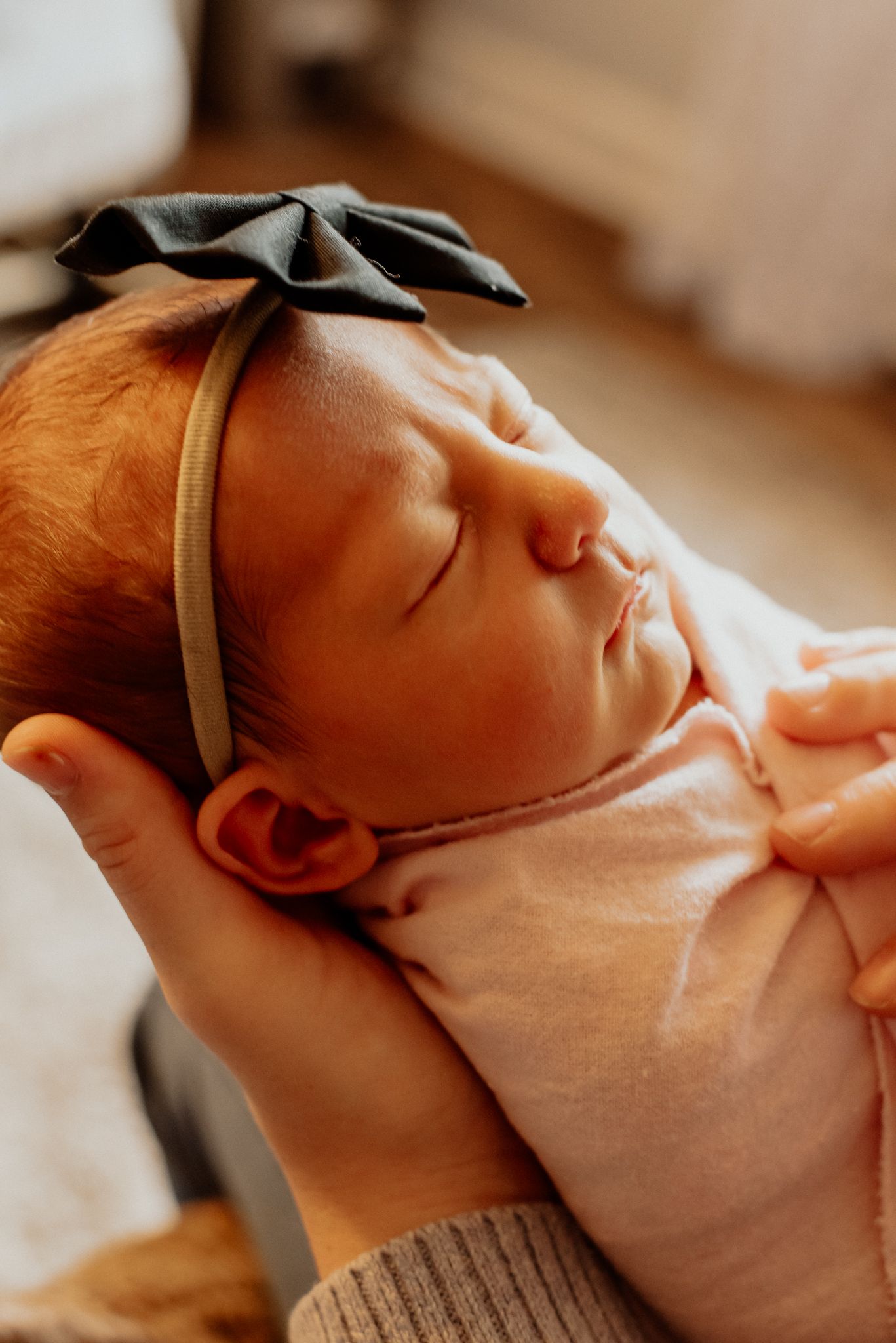 Newborn baby wearing pink outfit and gray bow headband sleeping peacefully.