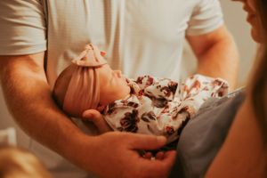 Newborn baby wearing floral outfit being cradled in arms in warm lighting.