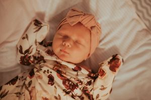 Sleeping newborn wearing pink headband and floral print outfit on white bedding.