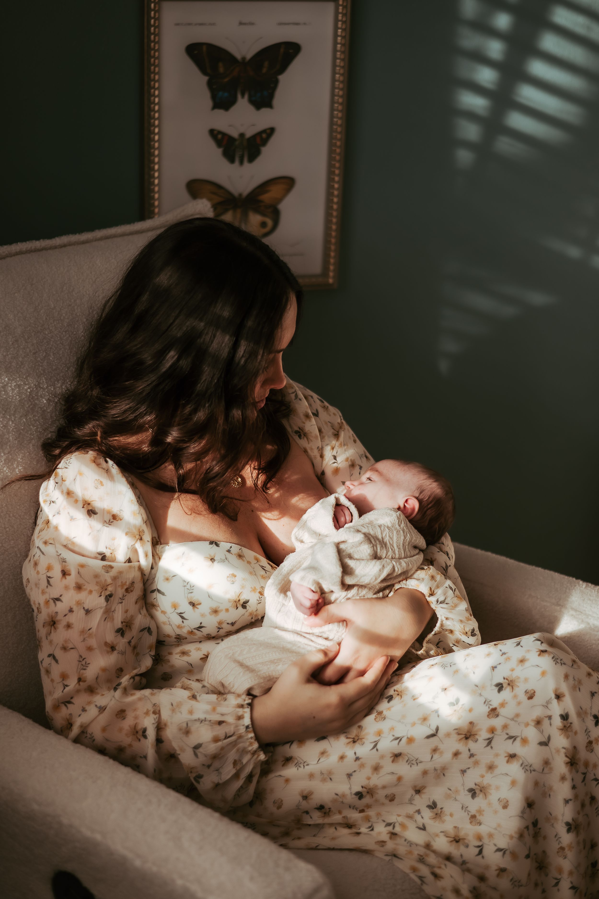 A tender moment captured as mother in floral dress cradles newborn baby in a chair beneath vintage butterfly art.