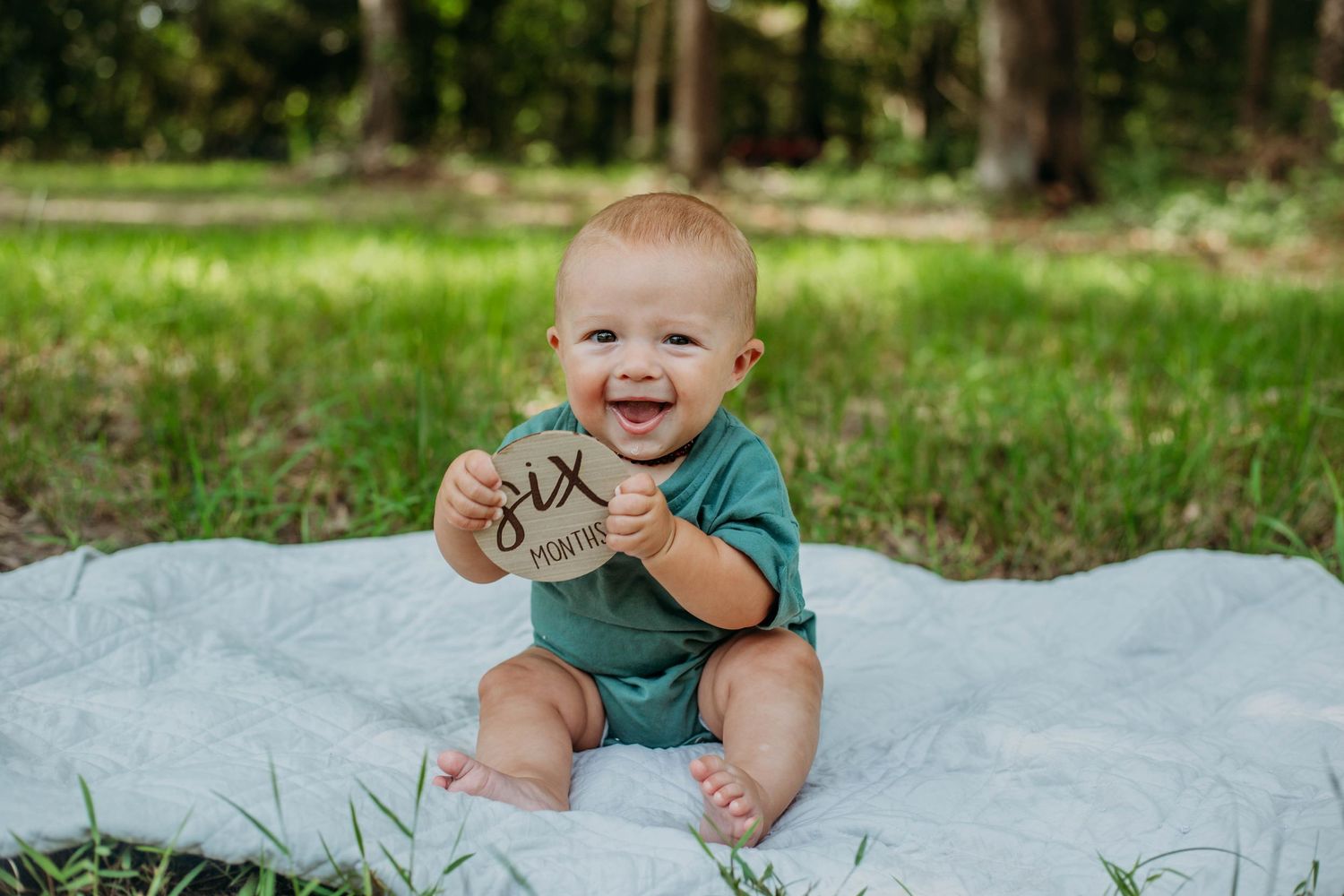 Happy baby in teal outfit sitting on white blanket outdoors holding pine cones.