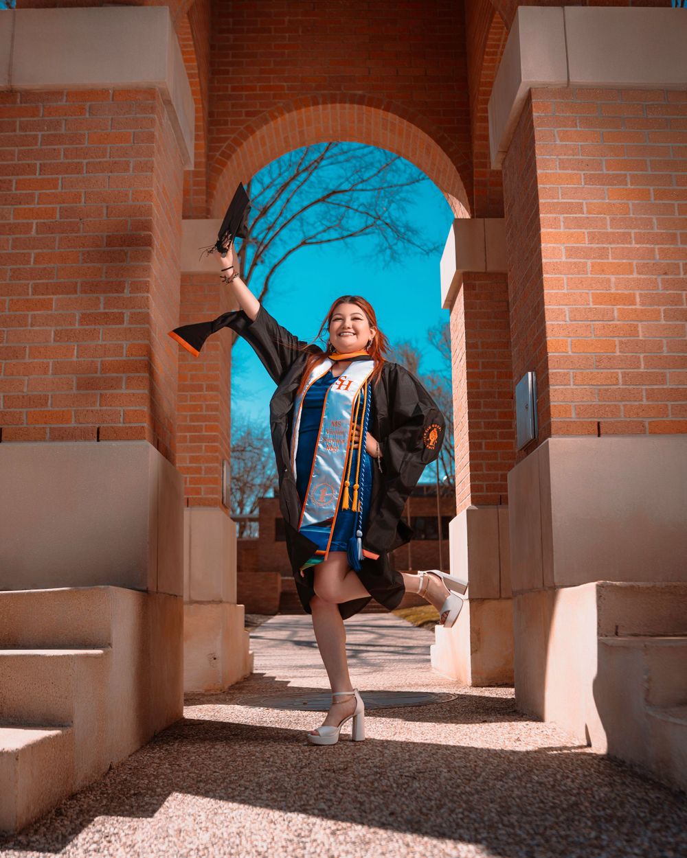 Graduate standing in brick archway wearing graduation regalia and heels against vibrant blue sky background.