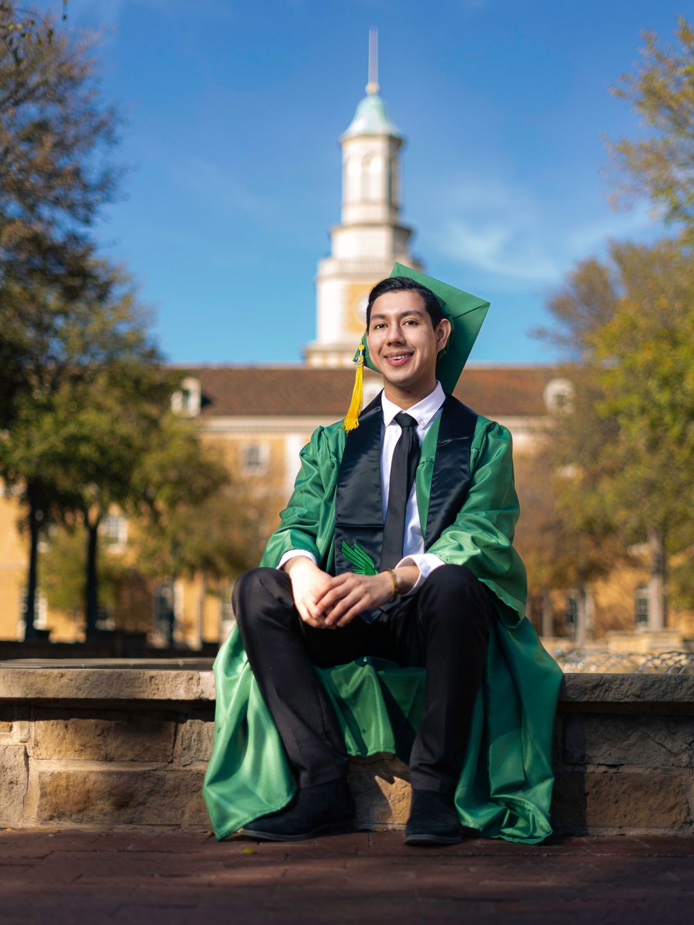 Graduate in green commencement robes sitting on wall with church steeple in background.
