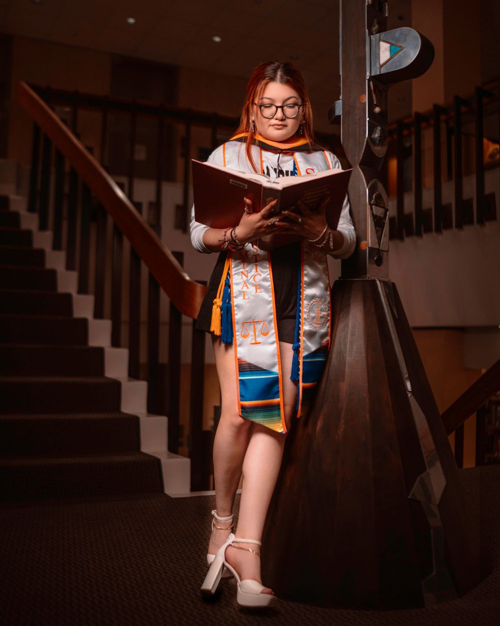 A reader in glasses and a decorative stole stands on wooden stairs while reading from a book in moody lighting.