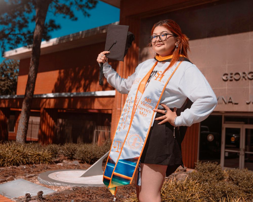 Outdoor graduation photo with orange graduation stole and white hoodie against brick building on sunny day.