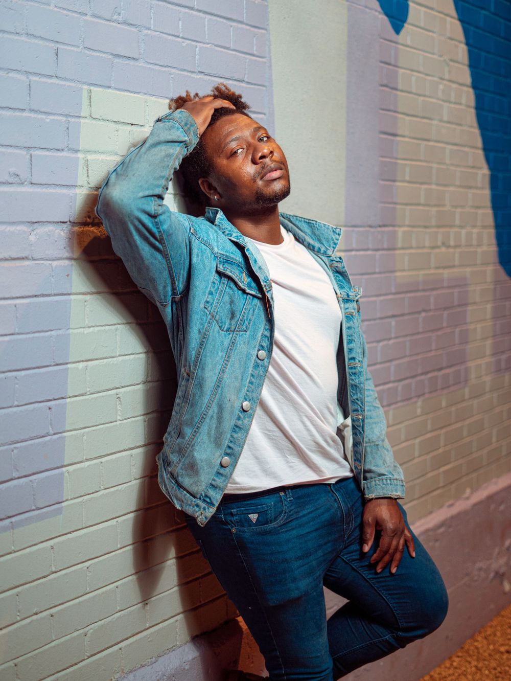 Casual portrait of person in denim jacket leaning against white brick wall.
