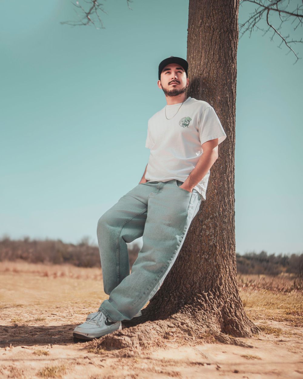 Someone in casual wear with a baseball cap leans against a tree trunk in a field with turquoise sky background.