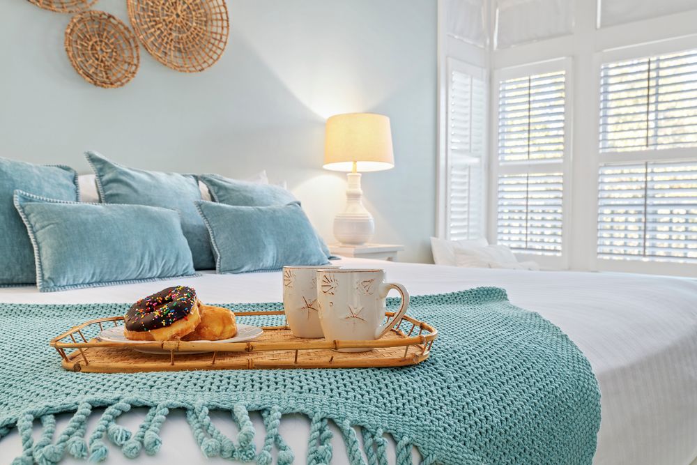 Cozy bedroom with turquoise bedding, white shuttered windows, and breakfast tray featuring toast and coffee.