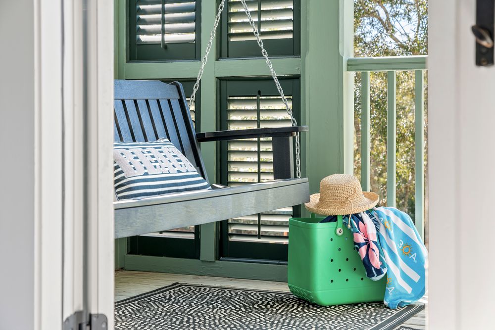 A cozy porch swing with blue cushions and green shutters features a woven basket and straw hat creating a relaxing outdoor space.