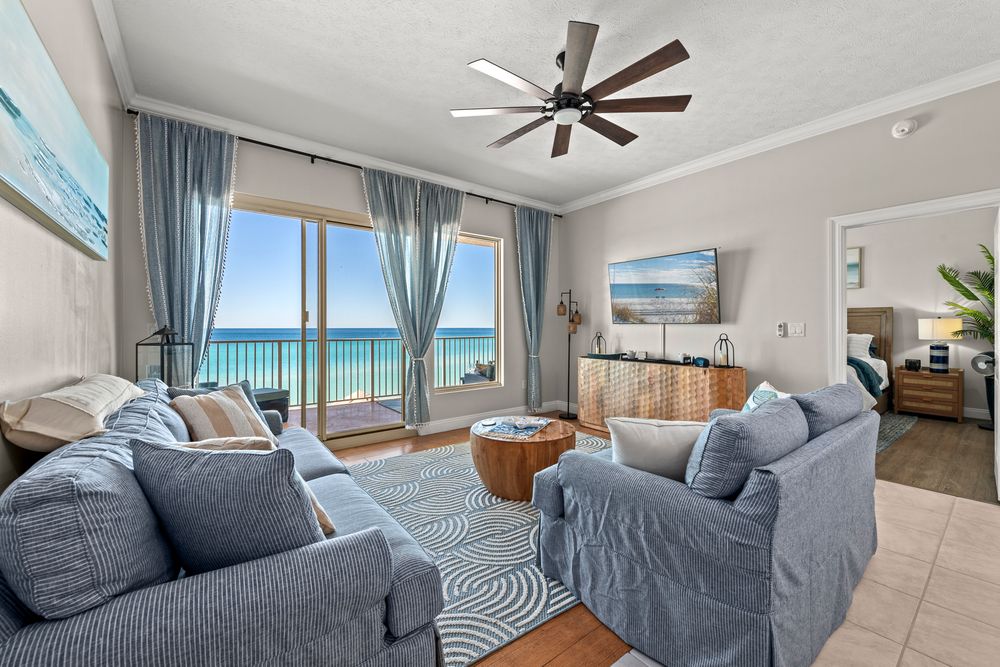 Coastal living room with gray sectional sofa, ceiling fan, and panoramic ocean views through large windows.