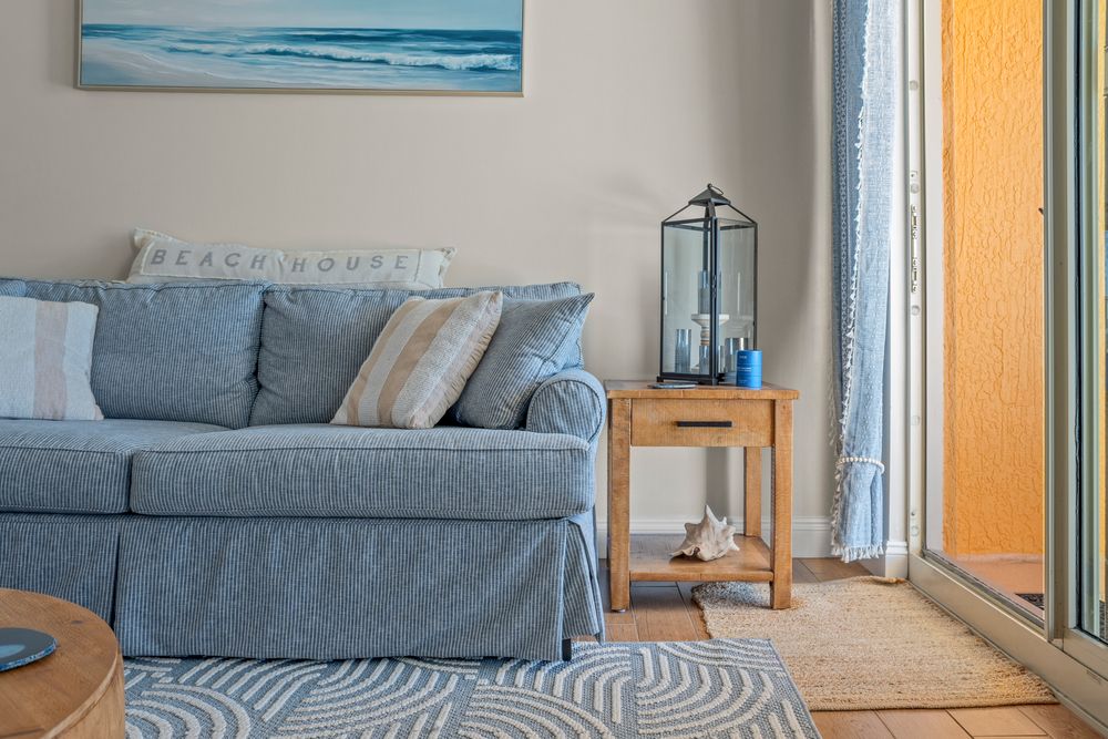 Coastal living room features a blue sofa, wooden side table with lantern, and beachy wall art above.