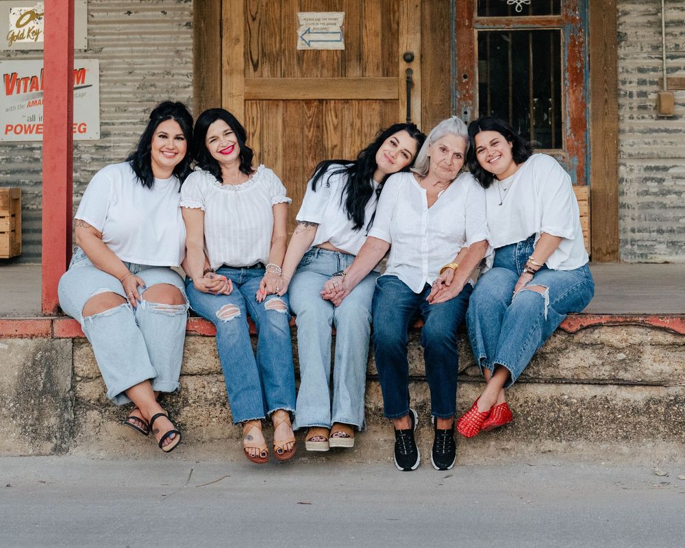 Group of women in matching white shirts and denim pose together on wooden storefront steps.