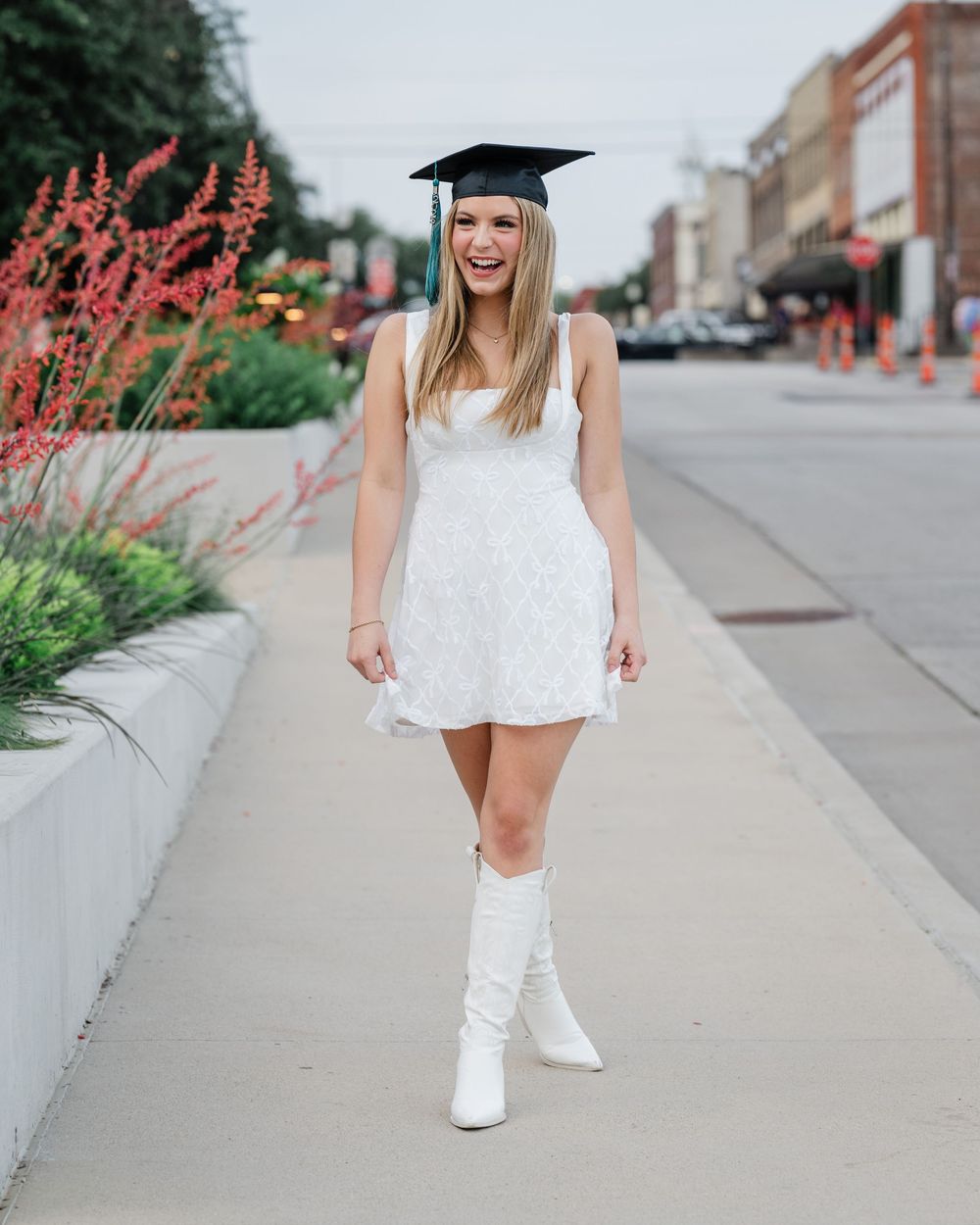 Graduate in white dress and cap poses on city sidewalk with boots and pink flowers in background.