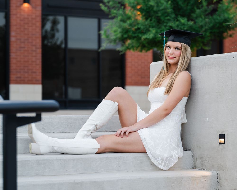 A graduation portrait on concrete steps featuring a white lace dress and matching sneakers against a brick building backdrop.
