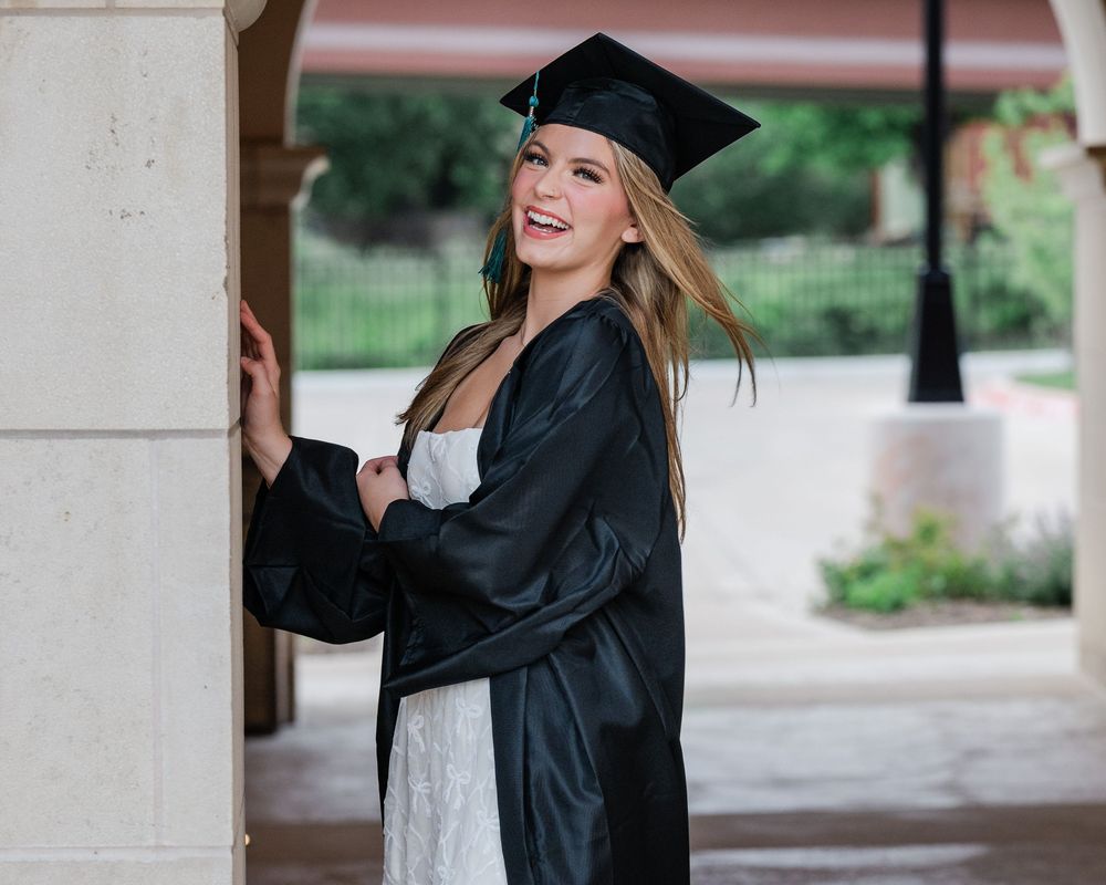 Graduate in black cap and gown stands joyfully by column during graduation photoshoot in outdoor setting.