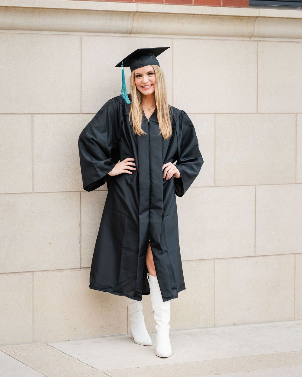 Graduate in black cap and gown poses confidently in white boots against neutral wall during graduation photoshoot.