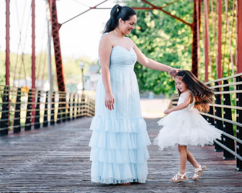 Woman in flowing blue dress and child in white tulle dress twirling together on wooden bridge.