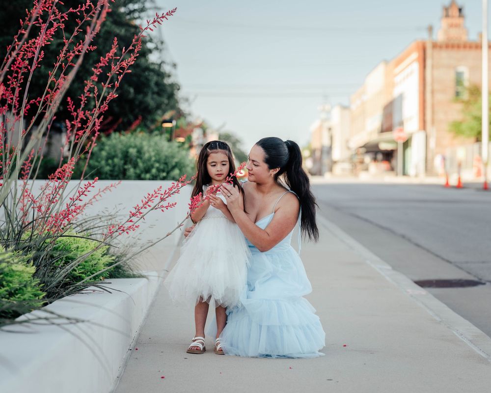 Parent kneeling beside child in white dress near blooming red flowers along city sidewalk.