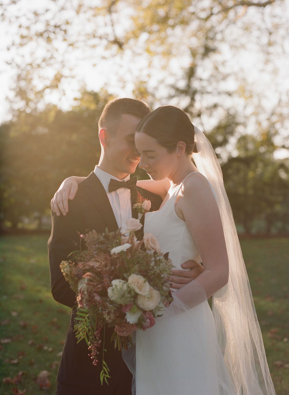 Bride and groom share a romantic golden hour moment outdoors, with bride holding a lush autumn bouquet.