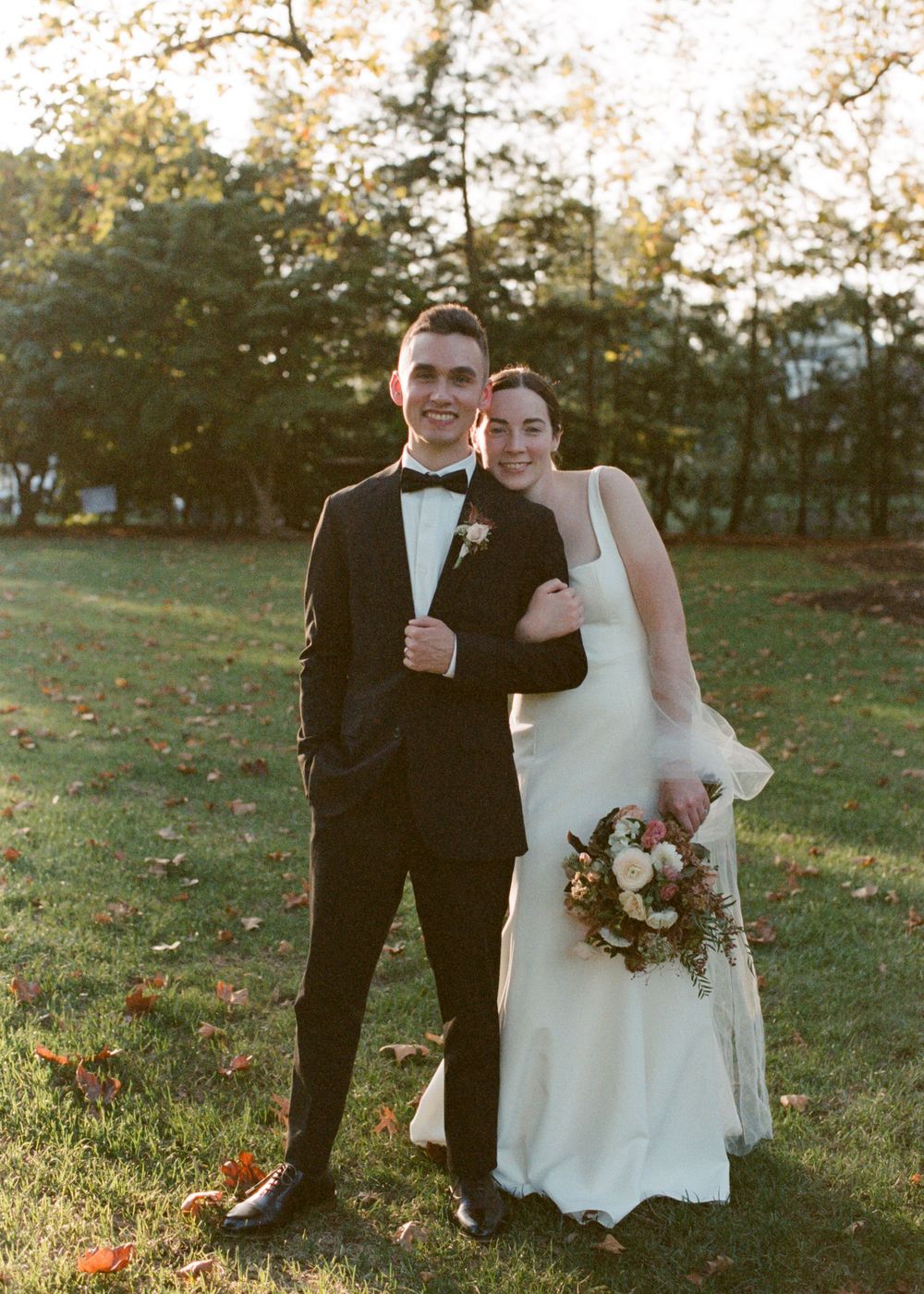 Couple poses in golden hour light on green lawn, groom in black tuxedo, bride in white gown holding floral bouquet.