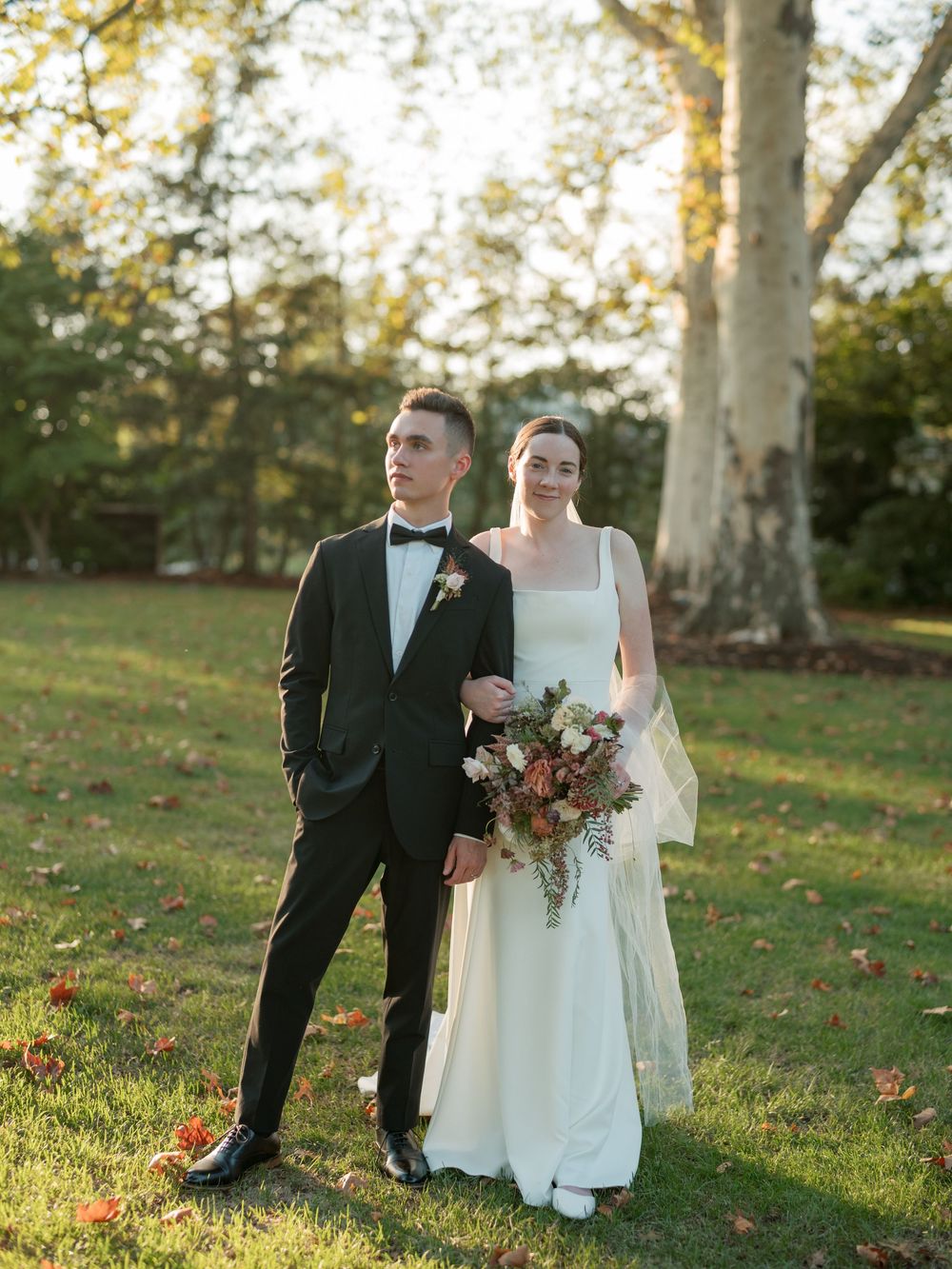 Elegant bride and groom posing outdoors in golden hour light, she holds a fall bouquet, he wears a black tuxedo.