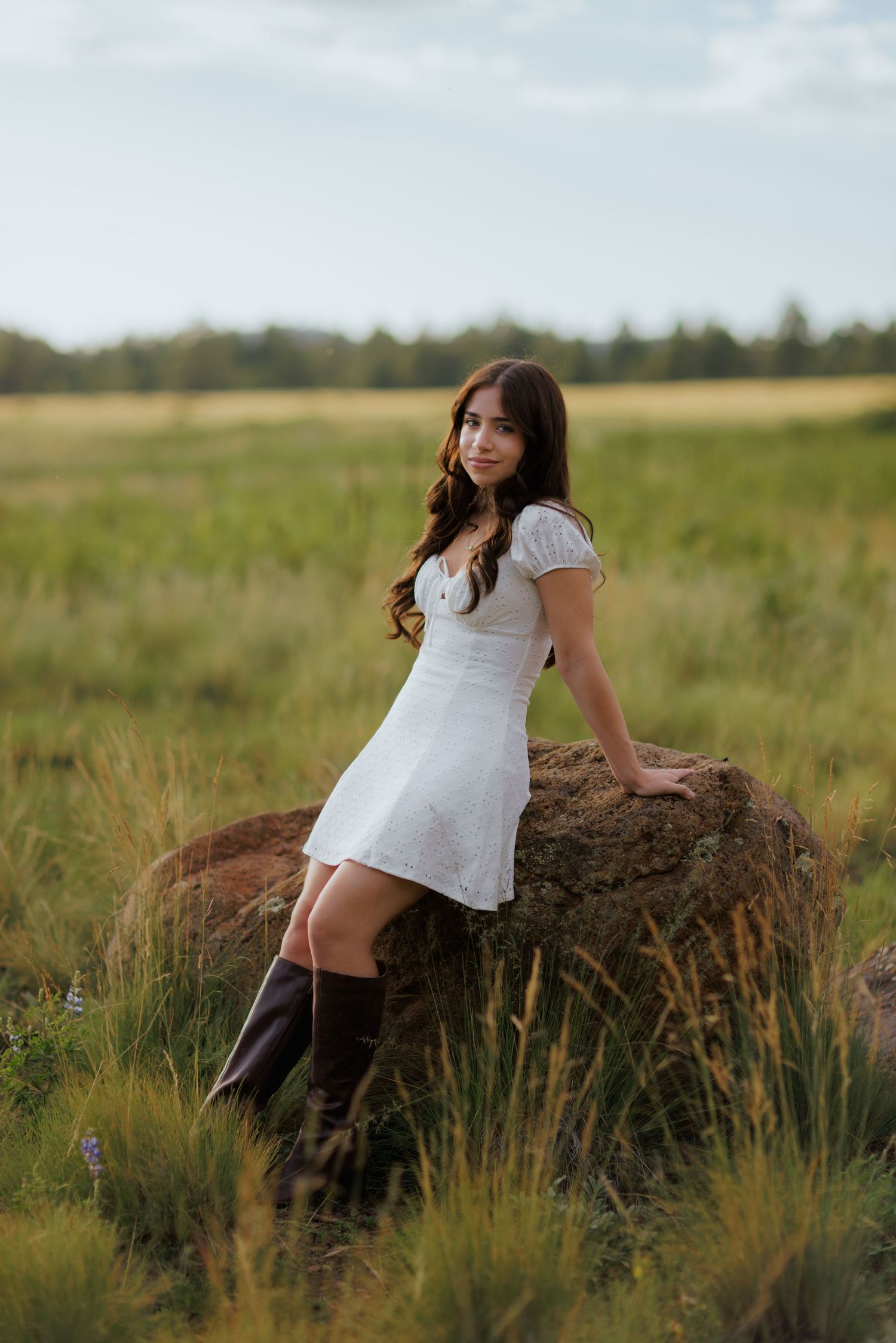 A brunette girl in a white dress and tall brown boots leans against a large rock in a green meadow.
