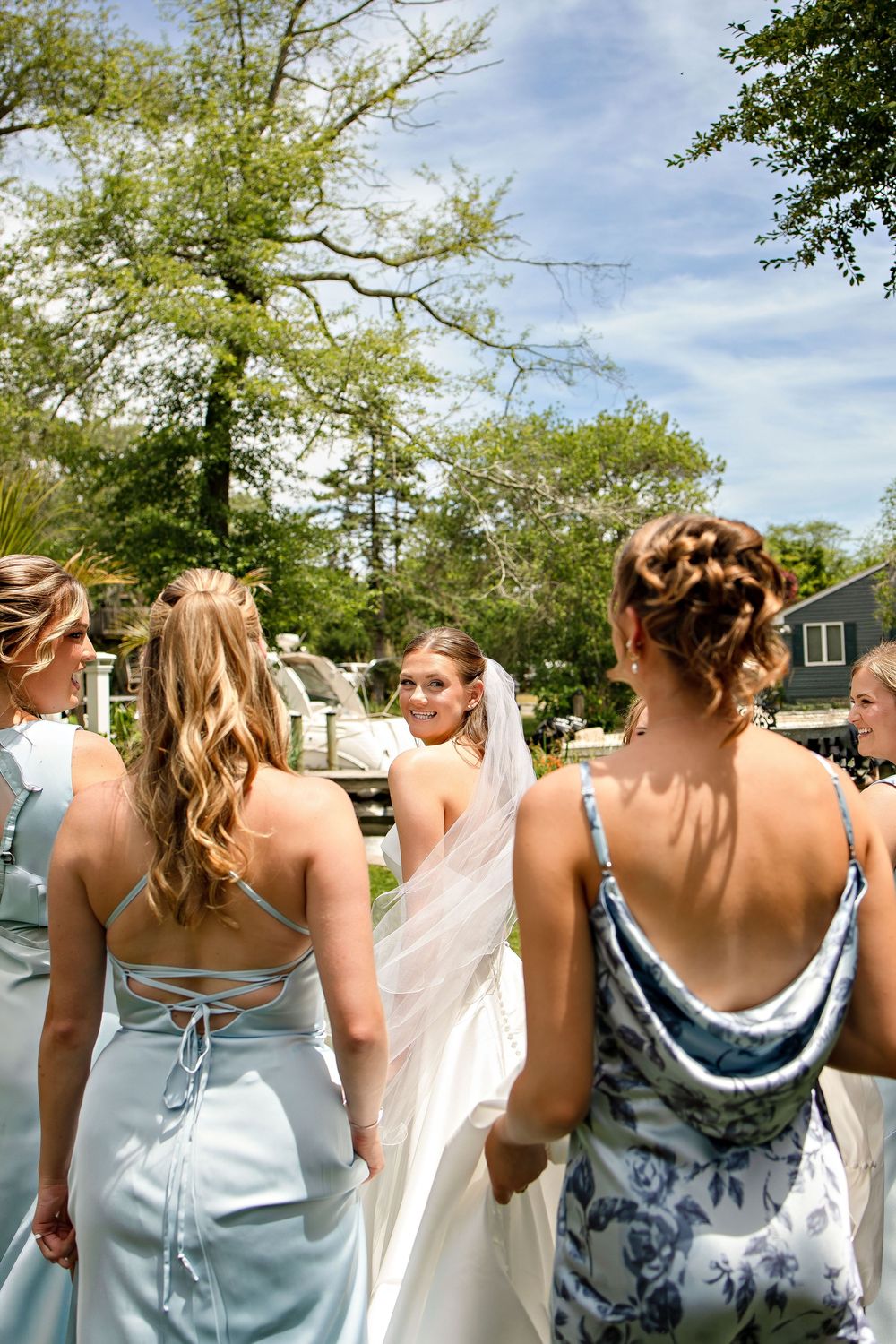 Bride in white gown and veil walks with bridesmaids in light blue dresses outdoors on a sunny wedding day.
