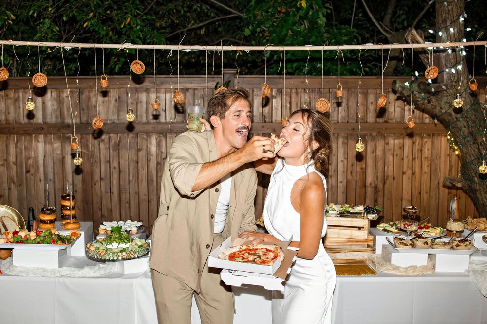 A joyful couple feeds each other cake at an outdoor wedding reception with string lights and wooden fence backdrop.