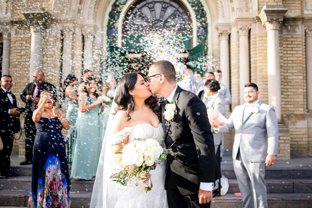 Wedding couple shares kiss under confetti shower at church entrance.