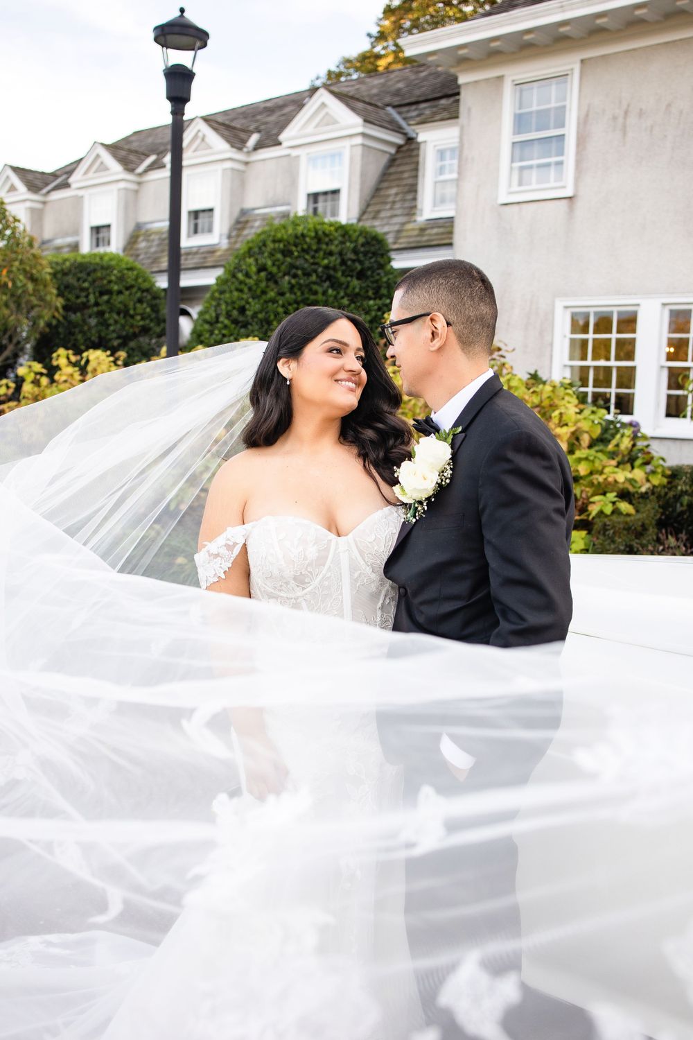 Wedding couple poses with flowing veil against white house exterior.