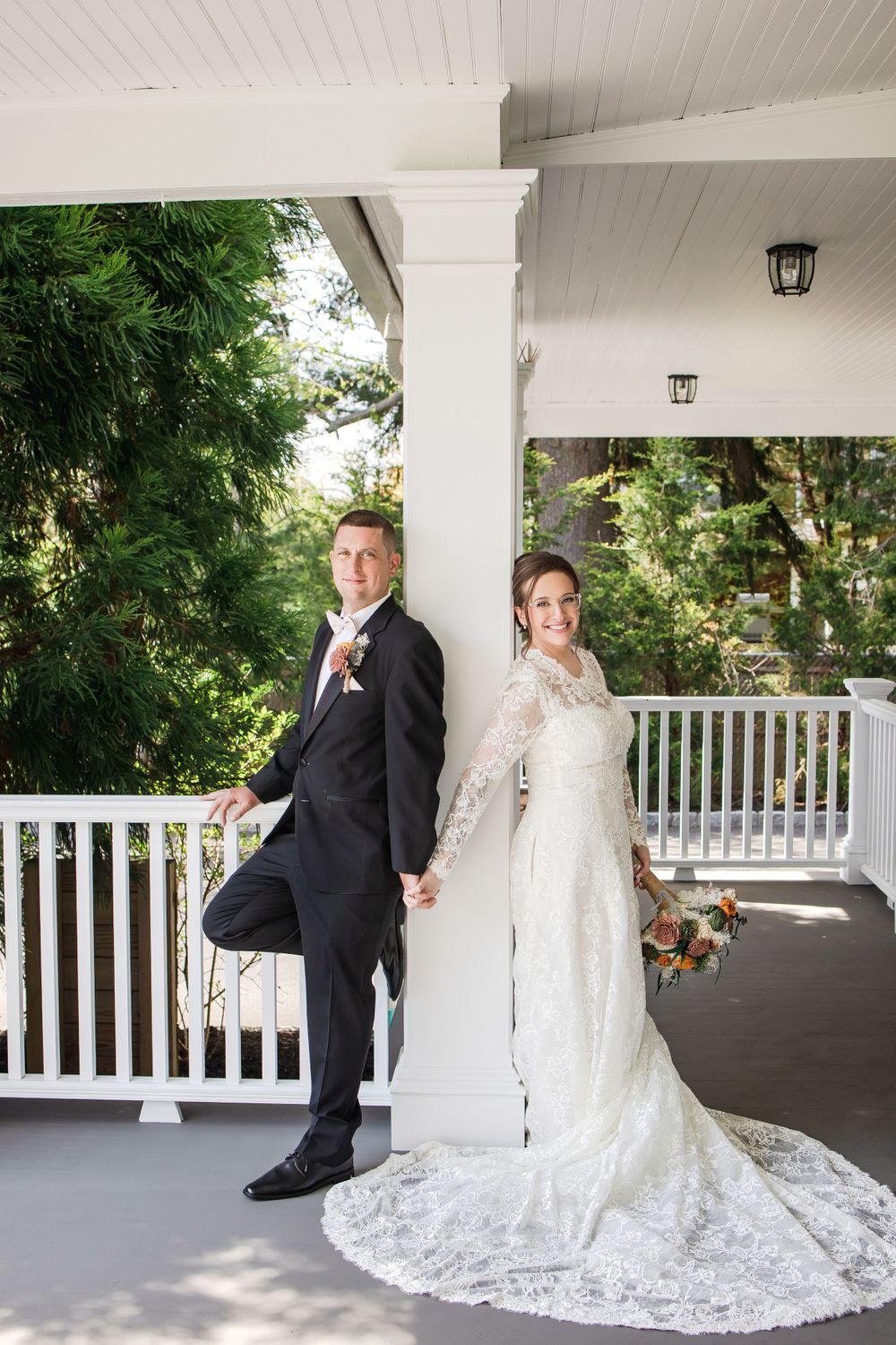 Elegant wedding portrait on a white porch with lush greenery in the background.